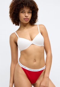 Biały stanik wypełniony z cienkimi ramiączkami, w połączeniu z czerwonym stringami z białą anteczką z logo "TOMMY HILFIGER". Gładki materiał, minimalistyczny design.