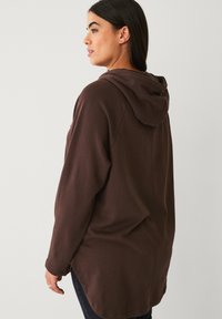 Brown hooded sweatshirt with a relaxed fit, long sleeves, textured fabric, and a longer back hem. Features a simple design without embellishments.