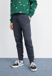 Graue Nike-Sweatpants mit einem elastischen Bund und Taschen, kombiniert mit schwarz-weißen karierten Vans-Sneakern auf einem blauen Teppich.