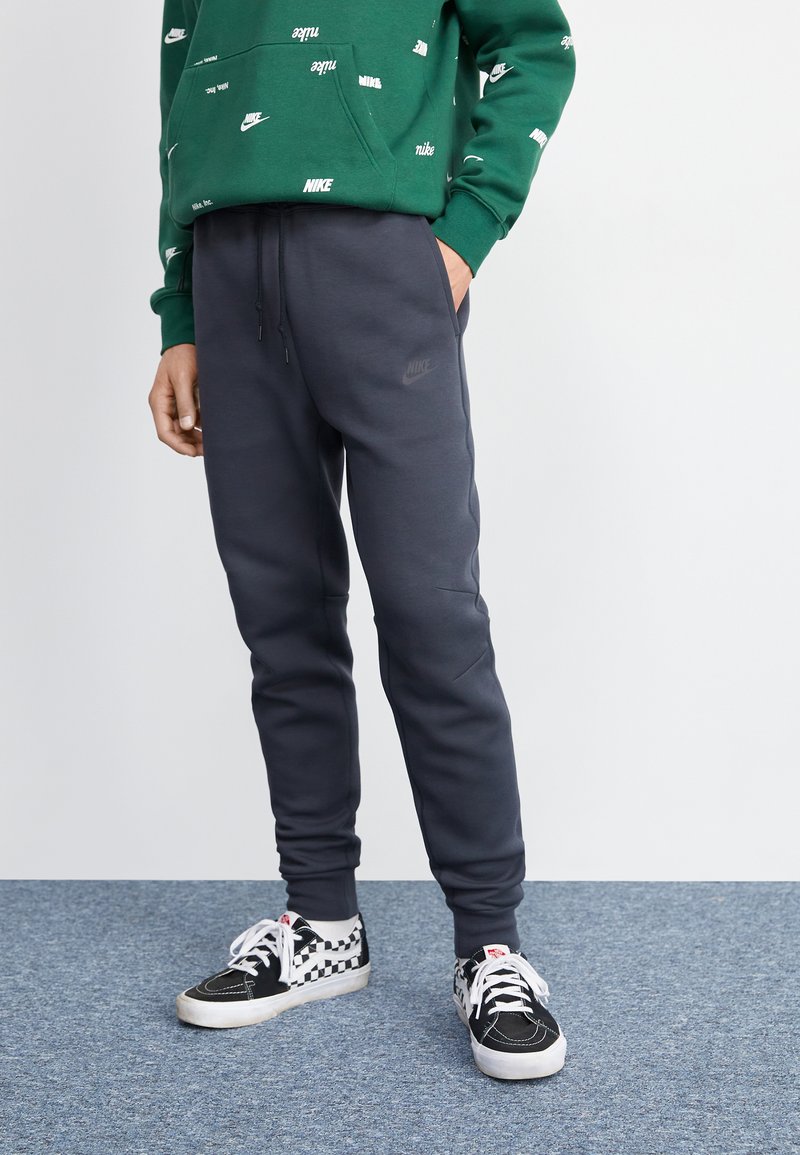 Graue Nike-Sweatpants mit einem elastischen Bund und Taschen, kombiniert mit schwarz-weißen karierten Vans-Sneakern auf einem blauen Teppich.