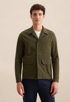 Giacca camicia verde oliva con maniche lunghe, due tasche frontali e chiusura a bottone. Il tessuto appare liscio e strutturato.