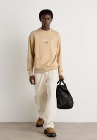 Beige cotton sweatshirt with a small rectangular graphic, paired with white loose-fit pants and brown slip-on shoes. Black bag in hand.