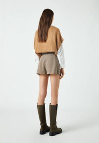 Poncho de punto marrón sobre una camisa blanca, combinado con pantalones cortos de pata de gallo marrones y botas verdes hasta la rodilla, vista desde la parte posterior.