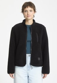 Giacca in pile nera con zip frontale, tasche laterali e un logo ricamato discreto. Presenta un tessuto testurizzato con una finitura morbida e polsini elastici.