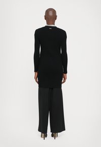 Black ribbed long-sleeve top with a slim fit, featuring a round neckline and a gold accent at the back, styled with black wide-leg trousers.