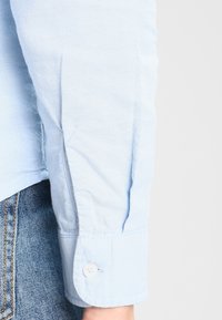 Ljusblå skjorta med knappar och vävd textur, med en uppvikt ärm och en ensam vit knapp på ärmen.