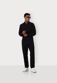 Schwarzes Langarmhemd mit Knöpfen, kombiniert mit passenden Hosen und weißen Sneakern; Sonnenbrille vervollständigt den minimalistischen Look.