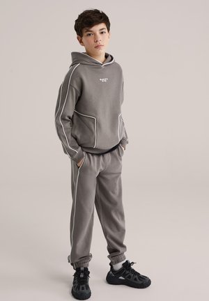 Hanorac gri și pantaloni de trening cu detalii albe, confecționați dintr-un material moale. Tenisi negri completează ținuta. Design casual cu buzunare.