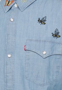 Light blue denim shirt featuring embroidered bees and butterflies. It has a chest pocket, snap buttons, and a fabric label on the side.