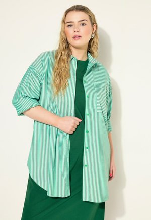 Camisa de rayas verdes y blancas de gran tamaño con mangas cortas, cierre frontal con botones y un solo bolsillo en el pecho. Combinada con un vestido verde sólido debajo.