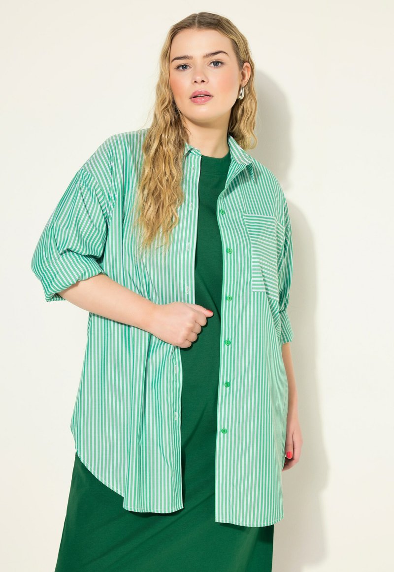 Camisa de rayas verdes y blancas de gran tamaño con mangas cortas, cierre frontal con botones y un solo bolsillo en el pecho. Combinada con un vestido verde sólido debajo.