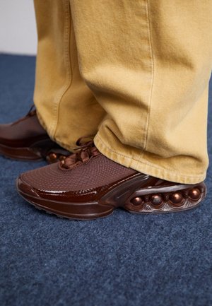 Personne portant des baskets marron avec un amorti sphérique unique et un pantalon jaune ample, debout sur un tapis bleu.
