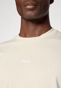 Kremfarget bomulls T-skjorte med rund hals og en hvit, kursiv logo "Rapha" på brystet. Glatt tekstur, tettsittende design.