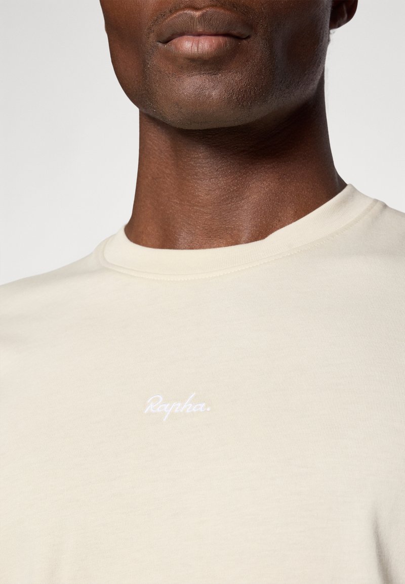 Cremfarbiges Baumwoll-T-Shirt mit Rundhalsausschnitt und einem weißen, geschwungenen Logo "Rapha" auf der Brust. Glatte Textur, taillierte Passform.