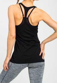Kvinde iført en sort sports tanktop med stropper og grå teksturerede leggings, stående med den ene hånd på hoften, set fra ryggen.