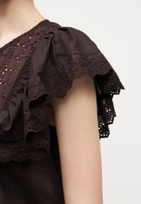 Αμάνικη μπλούζα σκούρου καφέ χρώματος με στρωμένες δαντελένιες βολάν στα μανίκια και κεντήματα με λουλούδια σε ρυθμό οπής στον ώμο και το άνω μέρος του στήθους.