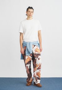 Fiatal személy fonott hajjal, fehér pólót visel, kék pulóver van a derekára kötve, és nagy lófej mintás nadrág van rajta.