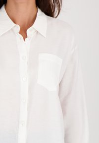 Vrouw in een witte overhemden met knopen, voorzien van een borstzak en een omgeslagen kraag, tegen een effen lichte achtergrond.
