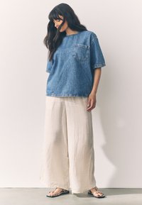 Top a maniche corte in denim blu con taschino sul petto, abbinato a pantaloni bianchi a gamba larga in lino e sandali bassi, su uno sfondo neutro.