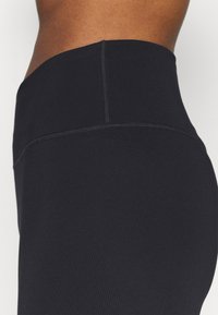 Gros plan sur la taille d'une personne portant des leggings noirs taille haute avec des coutures visibles sur un fond uni.