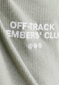 Zöld, textúrált anyag, rácsos mintázattal, fehér hímzett szöveggel: "OFF-TRACK EMBERS' CLUB" és az "OMC" logóval.