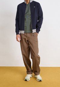 Giacca zip con colletto a scacchi in blu navy, t-shirt verde, pantaloni marroni e sneakers chiare; taglio casual con texture e modelli a contrasto.