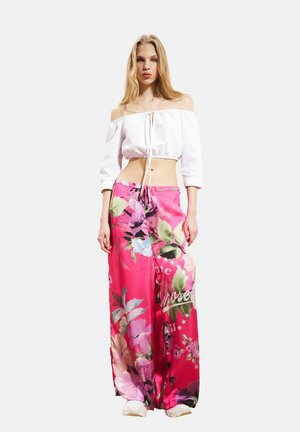 Γυναίκα στέκεται φορώντας λευκό crop top με ανοιχτούς ώμους και φωτεινά ροζ φαρδιά παντελόνια με μεγάλα λουλούδια και λευκά αθλητικά παπούτσια.