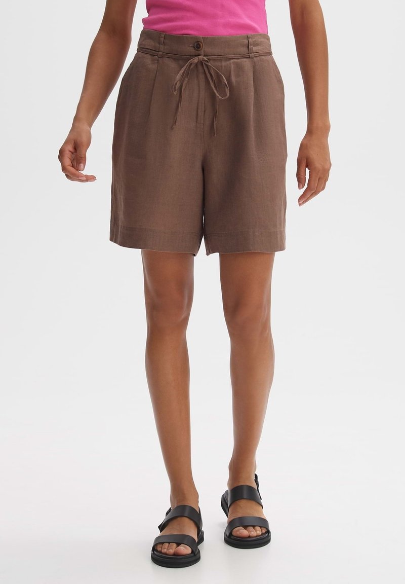 Shorts en lin marron avec devant plat, fermeture à bouton et cordon de serrage. Coupe décontractée et longueur jusqu'aux genoux, associés à des sandales noires.