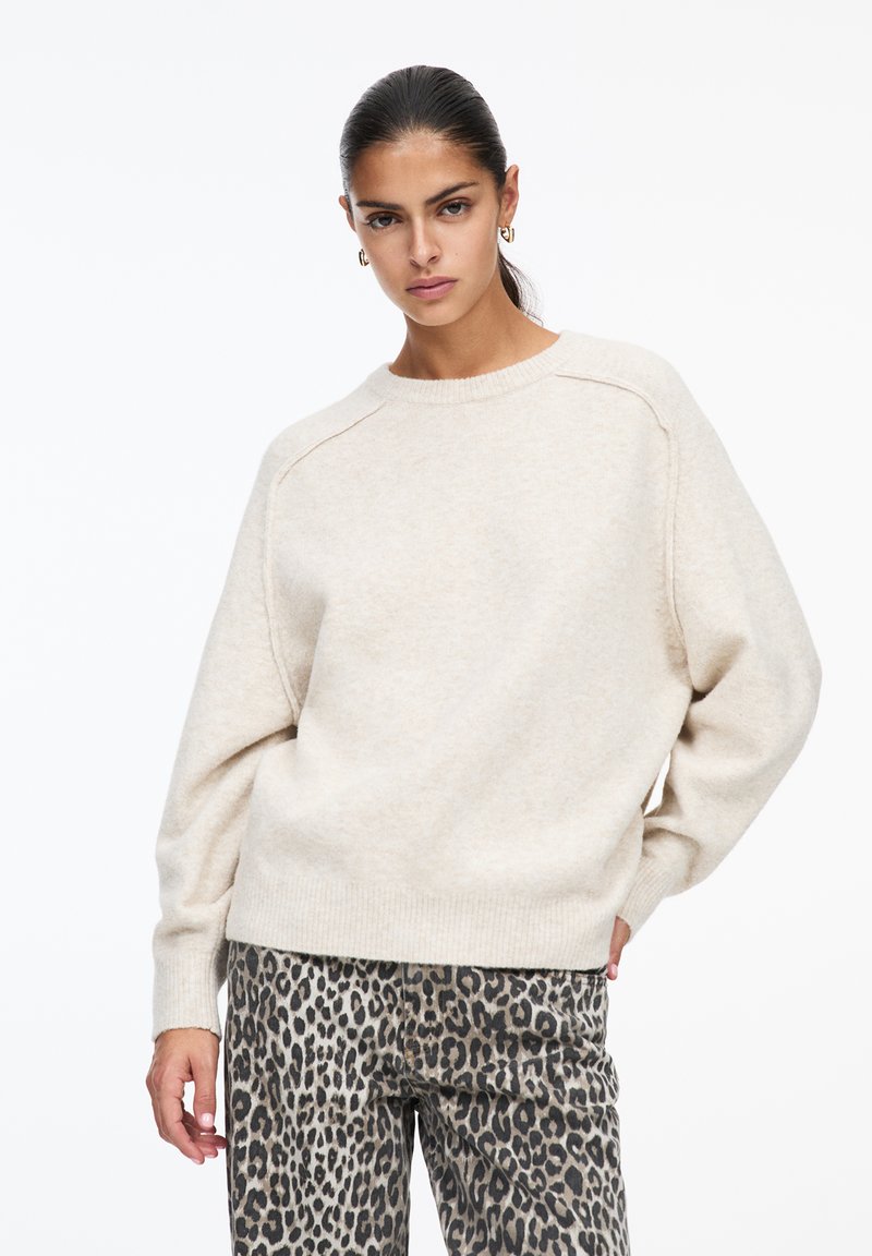 Pull en tricot couleur crème avec des manches longues, un col et un ourlet côtelés. Associé à un pantalon gris à imprimé léopard, affichant une texture douce.