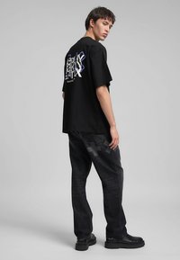 Maglietta nera oversize con stampa grafica sul retro, abbinata a jeans scuri con leggeri motivi a croce e stivali neri alla caviglia.