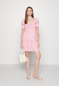 Vestido curto rosa com mangas bufantes, apresentando um padrão em ondas e uma saia em camadas. Combinado com uma bolsa branca e sandálias de salto com tiras.