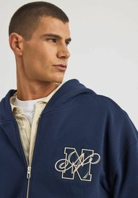 Granatowa bluza z zamkiem błyskawicznym z wyszywanym literą "M" w stylu kursywy. Gładka faktura, luźny krój i kaptur ze sznurkiem.