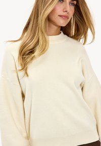 Pull en maille beige clair avec un col haut, des épaules tombantes et des finitions côtelées. Texture douce avec une coupe décontractée.