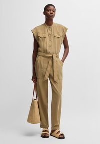 Khaki jumpsuit with a high neckline, button-up front, chest pockets, and a tied waist. Worn with a woven bag and sandals. Smooth texture.