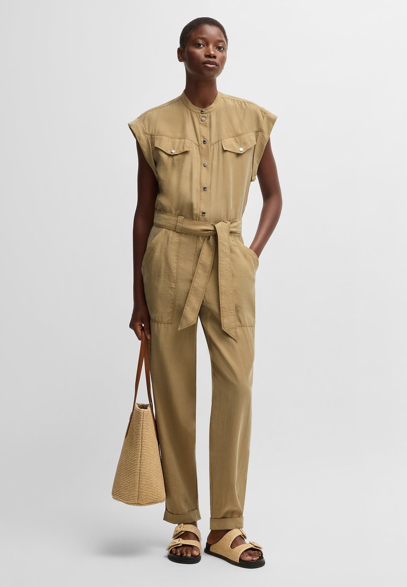 Khaki jumpsuit with a high neckline, button-up front, chest pockets, and a tied waist. Worn with a woven bag and sandals. Smooth texture.