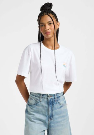 Weißes Baumwoll-T-Shirt mit kurzen Ärmeln und Rundhalsausschnitt, mit einem kleinen bunten Grafikdesign auf der linken Brust. Kombiniert mit hochtaillierten Jeans.