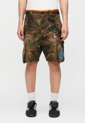 Muž nosiaci hnedé a zelené maskáčové cargo šortky s čiernymi cvokmi a modrou škvrnou od farby na pravej nohe, doplnené bielymi ponožkami a čiernymi teniskami.