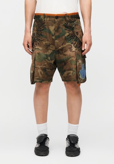 Homme portant un short cargo camouflage marron et vert avec des détails en clous noirs et une tache de peinture bleue sur la jambe droite, associé à des chaussettes blanches et des baskets noires.