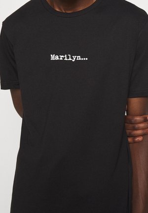 Zwart T-shirt met witte tekst "Marilyn..." gecentreerd op de borst, gedragen door een persoon met gekruiste armen gedeeltelijk zichtbaar.