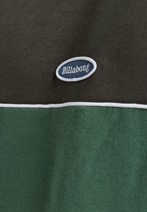 Close-up of a fabric with a dark green and black pattern, featuring a small blue and white Billabong logo.