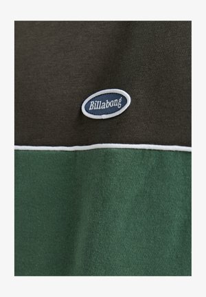 Close-up of a fabric with a dark green and black pattern, featuring a small blue and white Billabong logo.