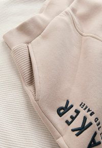 Manga de sudadera beige con puño acanalado y letras bordadas en negro "AKER" sobre un fondo de tejido suave y texturizado.