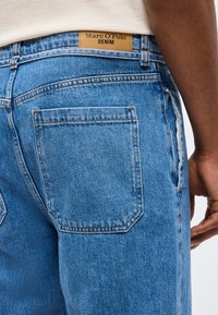 Primo piano di una persona che indossa jeans di jeans blu con un'etichetta marrone chiaro "Marc O’Polo DENIM" sopra la tasca posteriore e una camicia bianca infilata dentro.