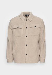 Beige fleece jacket with a collar, two chest pockets, and black buttons. Features a textured, soft surface and a straight cut.