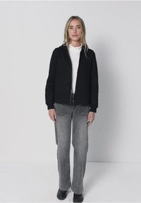 Zwarte jas met ritssluiting en een gestructureerd oppervlak, gedragen over een wit shirt, gecombineerd met lichtgrijze spijkerbroek en zwarte schoenen. Model staat tegen een effen achtergrond.