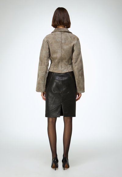 Skraćena smeđa jakna od antilopa s dugim rukavima, uparena s pripijenom crnom kožnom suknjom s prorezom na leđima i kontrastnim šavovima.