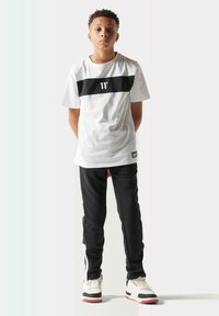 Camiseta blanca con raya horizontal negra, pantalones deportivos negros con rayas laterales blancas y zapatillas con acentos en negro, blanco y rojo.