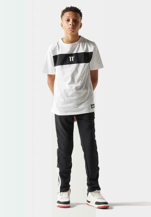 Camiseta blanca con raya horizontal negra, pantalones deportivos negros con rayas laterales blancas y zapatillas con acentos en negro, blanco y rojo.