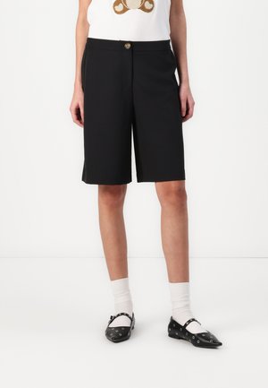 Shorts noirs arrivant au genou en tissu lisse, avec une fermeture à bouton à l'avant et des poches. Associés à un t-shirt graphique blanc et des chaussures noires.