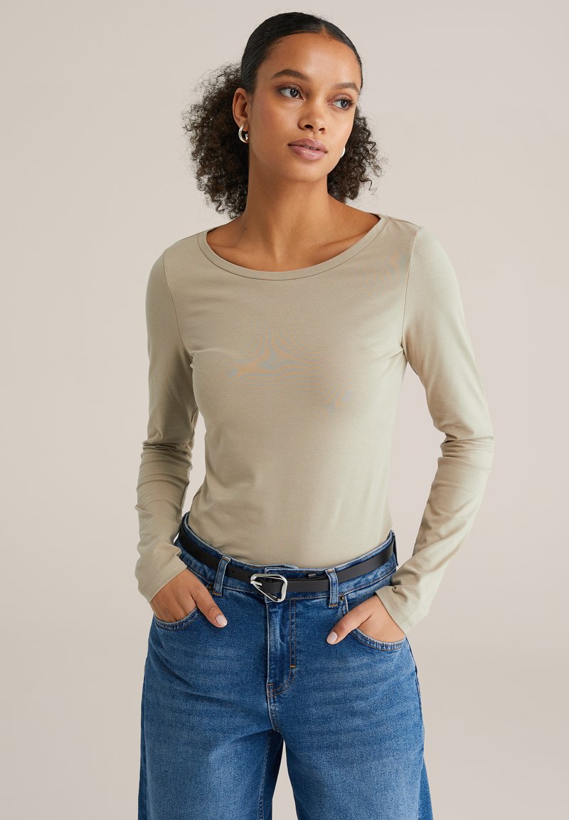 Šviesiai rudas ilgomis rankovėmis megztinis su apvalia iškirpte, derinamas su mėlynais aukšto liemens džinsais ir juodu diržu su sidabrine sagtimi.